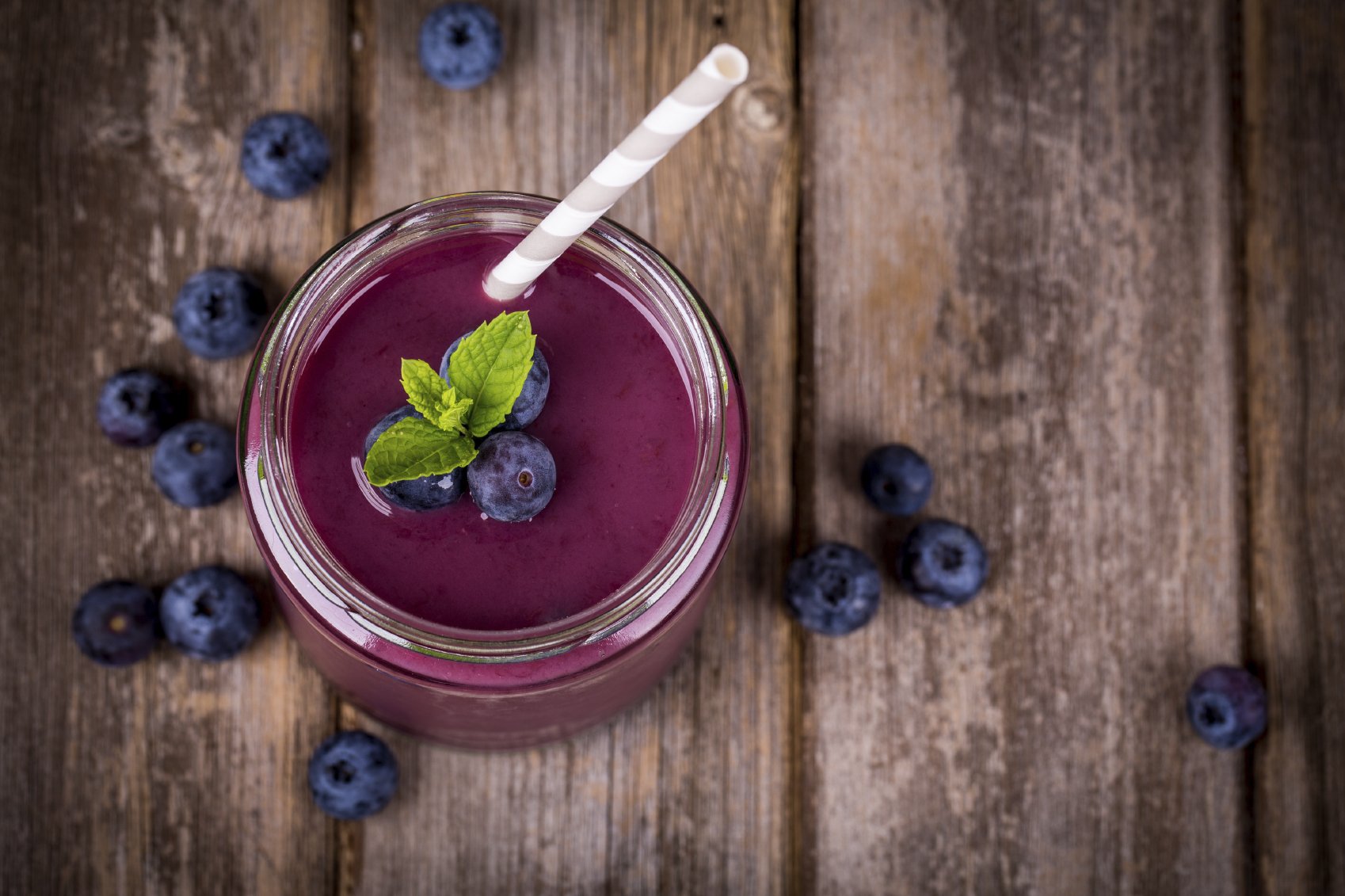 Blueberry smoothie in a glass jar with a straw and sprig of mint, over vintage wood table with fresh berries.