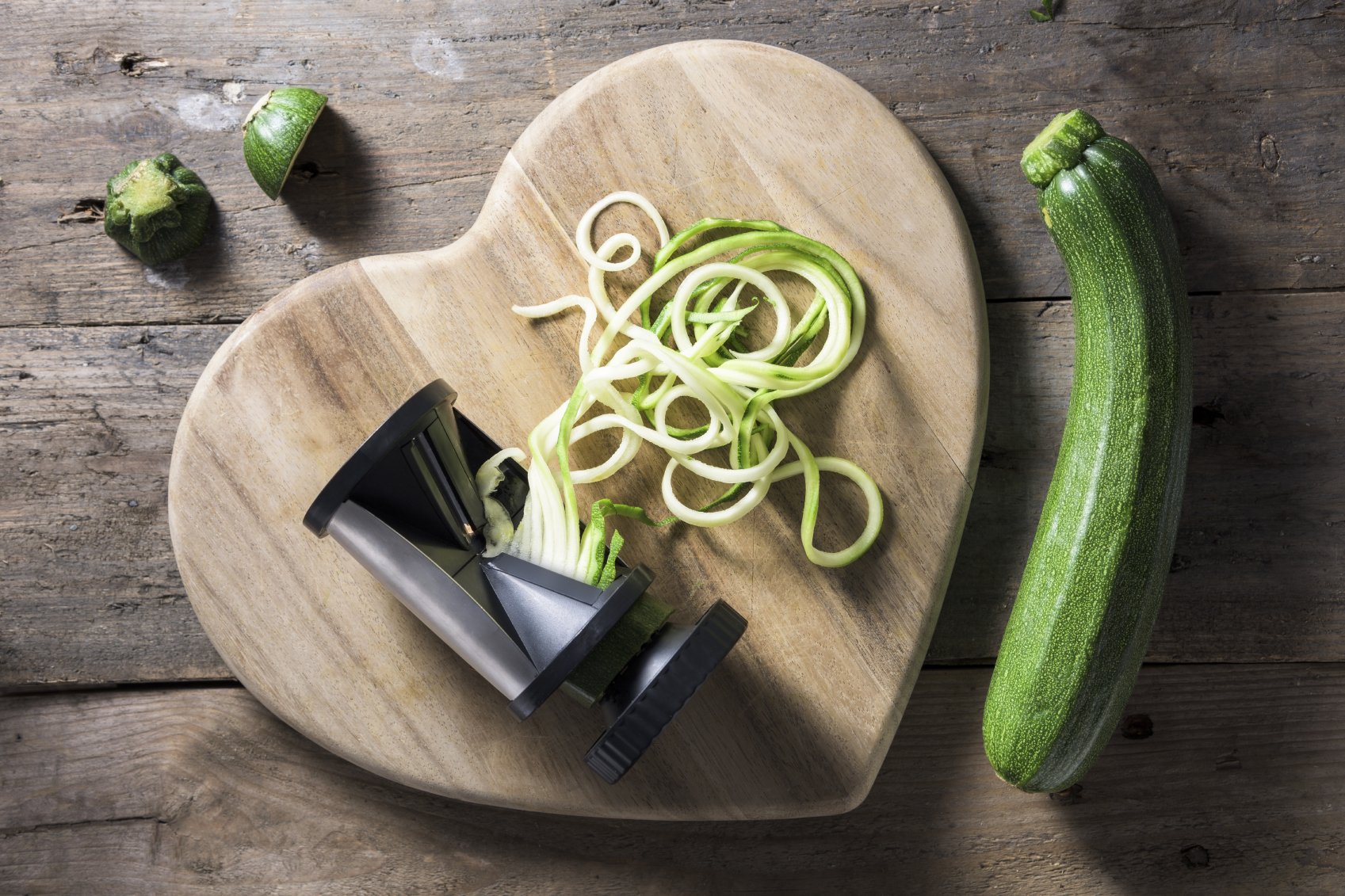 Freshly spiralised courgette twisting out of a spiraliser onto a heart shaped wooden chopping board. The courgette has been turned into long spaghetti shaped pieces, which look like noodles. A large green courgette is lying beside the chopping board with the top and tail of the courgette that has been used.