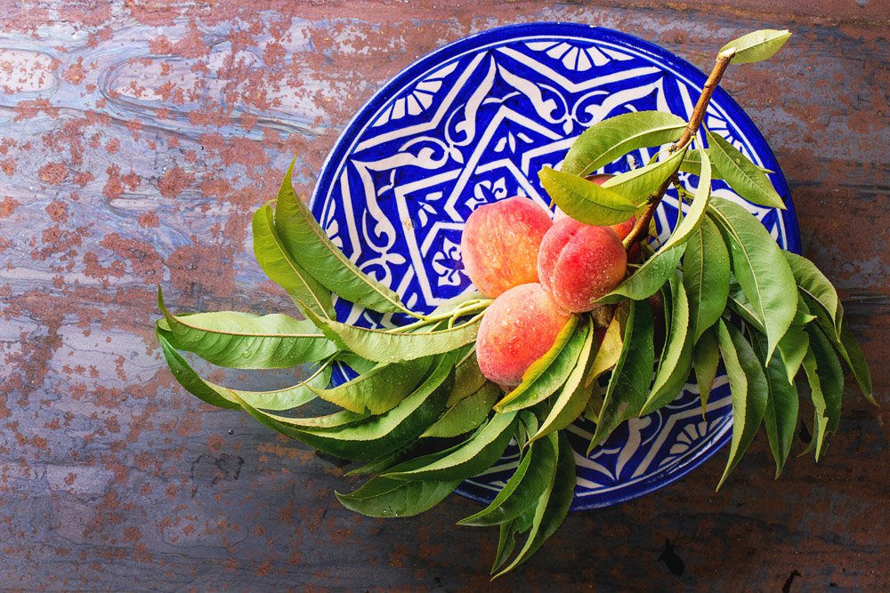 Peaches on branch with leaves on ornate blue plate over old metal background. Top view.