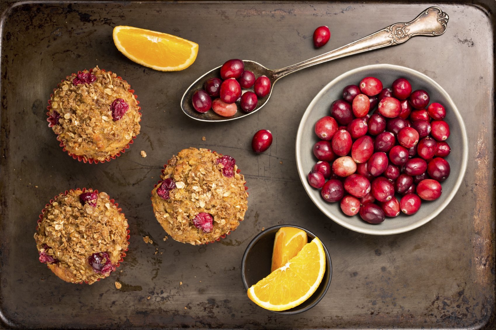 Freshly baked cranberry muffins on a rustic baking pan with cranberries and orange slices.
